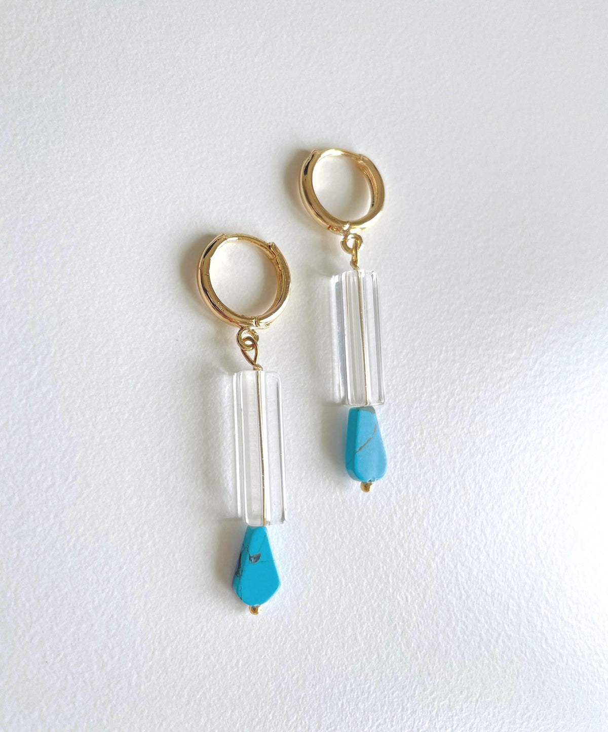 A pair of gold plated hoop drop earrings featuring clear cylindrical beads and bright turquoise blue teardrop beads, displayed on a clean white background.