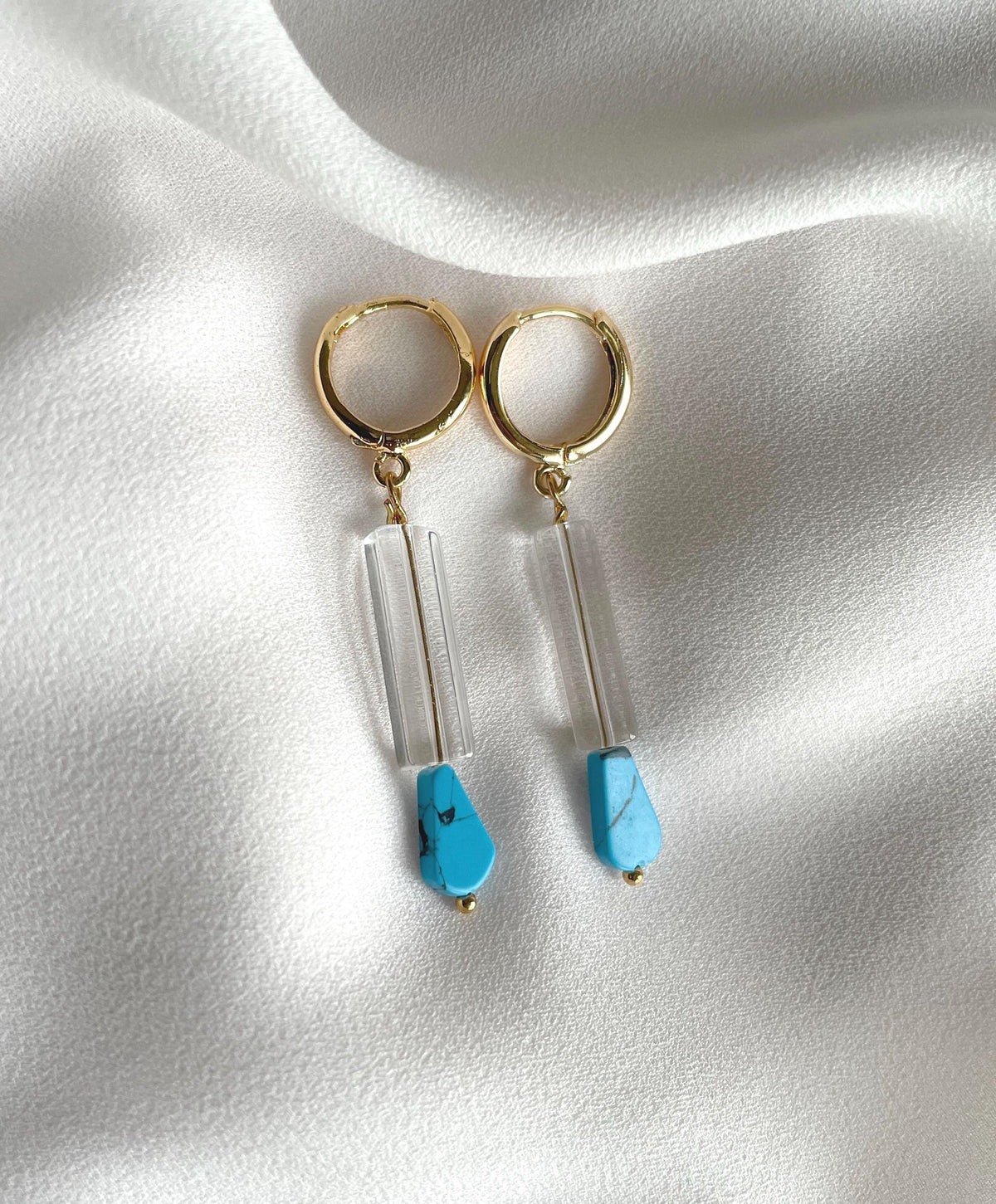 A pair of gold plated hoop dangle earrings with clear acrylic rectangular beads and light blue teardrop beads, displayed on a draped white fabric background.