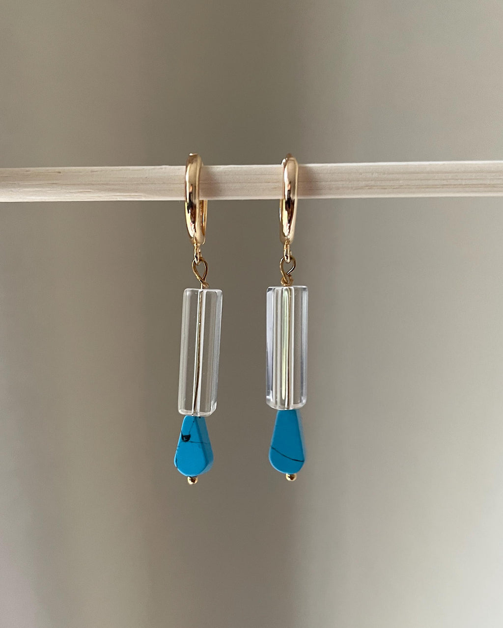 A pair of gold plated hoop dangle earrings with clear cylindrical beads and bright blue teardrop pendants, hanging from a light wooden rod against a soft gray background.