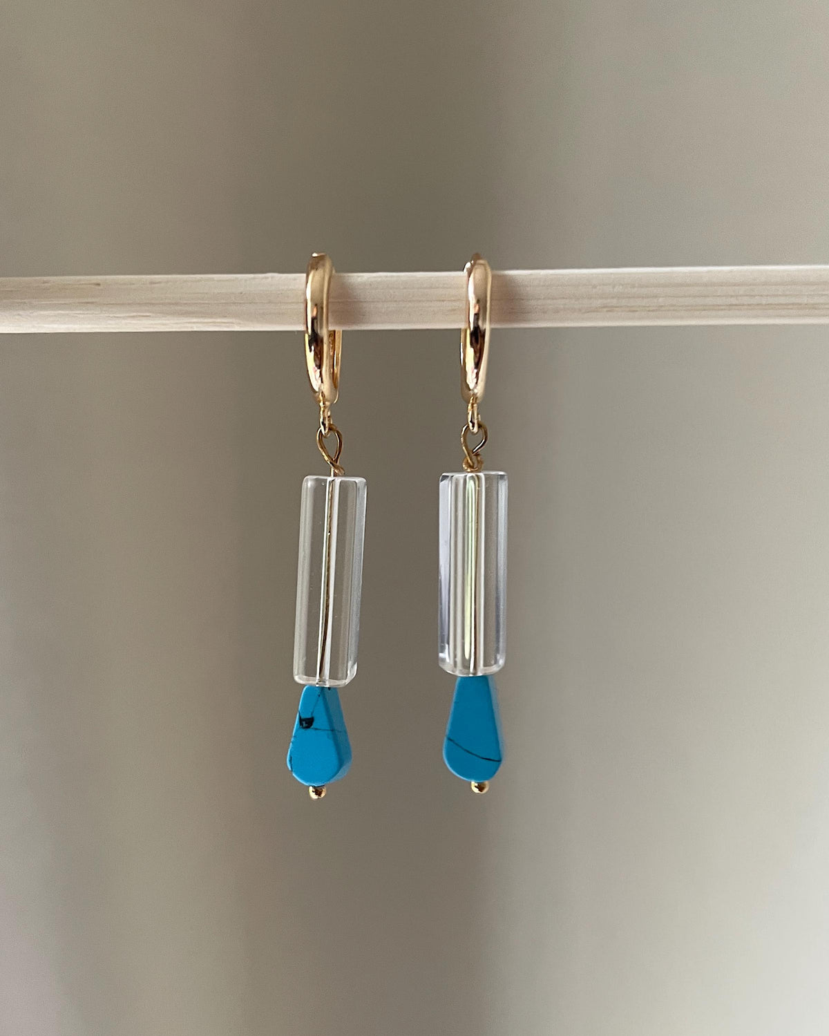 A pair of gold plated hoop dangle earrings with clear cylindrical beads and bright blue teardrop pendants, hanging from a light wooden rod against a soft gray background.