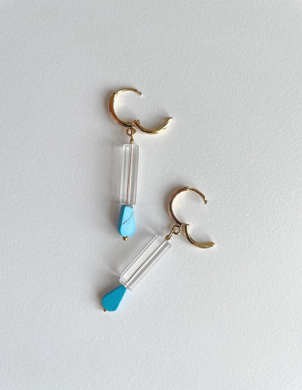 A pair of gold hoop dangle earrings with clear glass cylinder drops and small light blue teardrop-shaped accents, displayed on a light gray textured background.