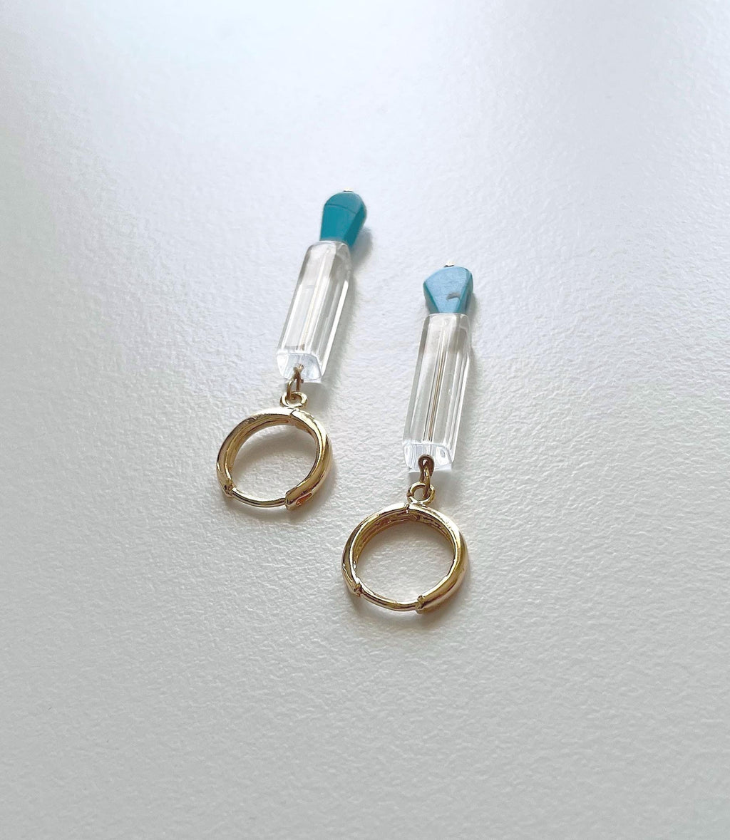 A pair of gold hoop earrings with clear faceted tube charms topped with light blue stone accents, displayed on a textured white surface.