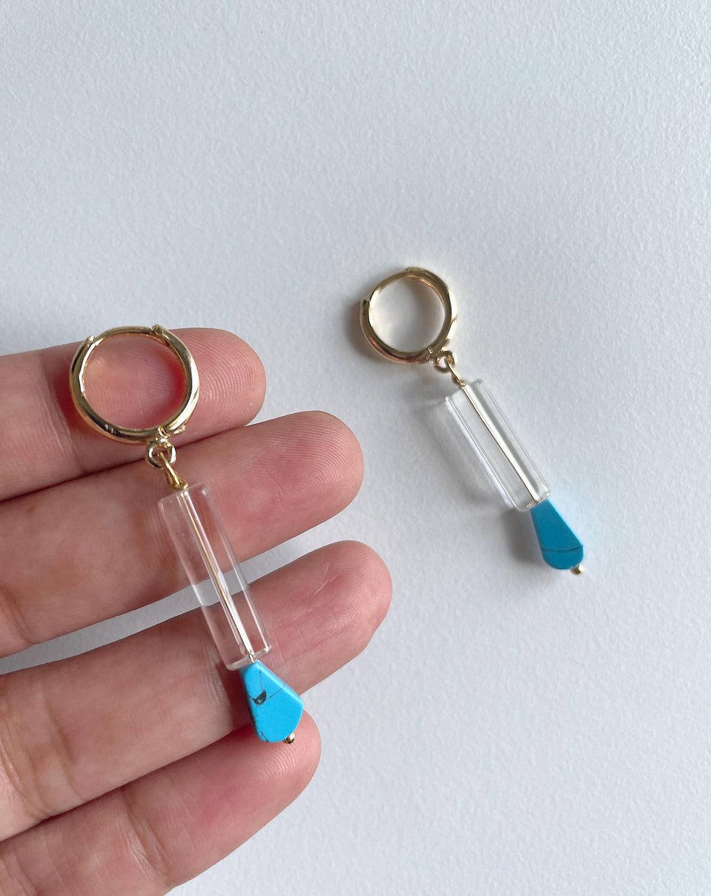 A pair of gold plated huggie hoop earrings with clear cylindrical beads and bright blue teardrop charms, shown with one keychain held on a person's fingers and the other resting on a white surface.