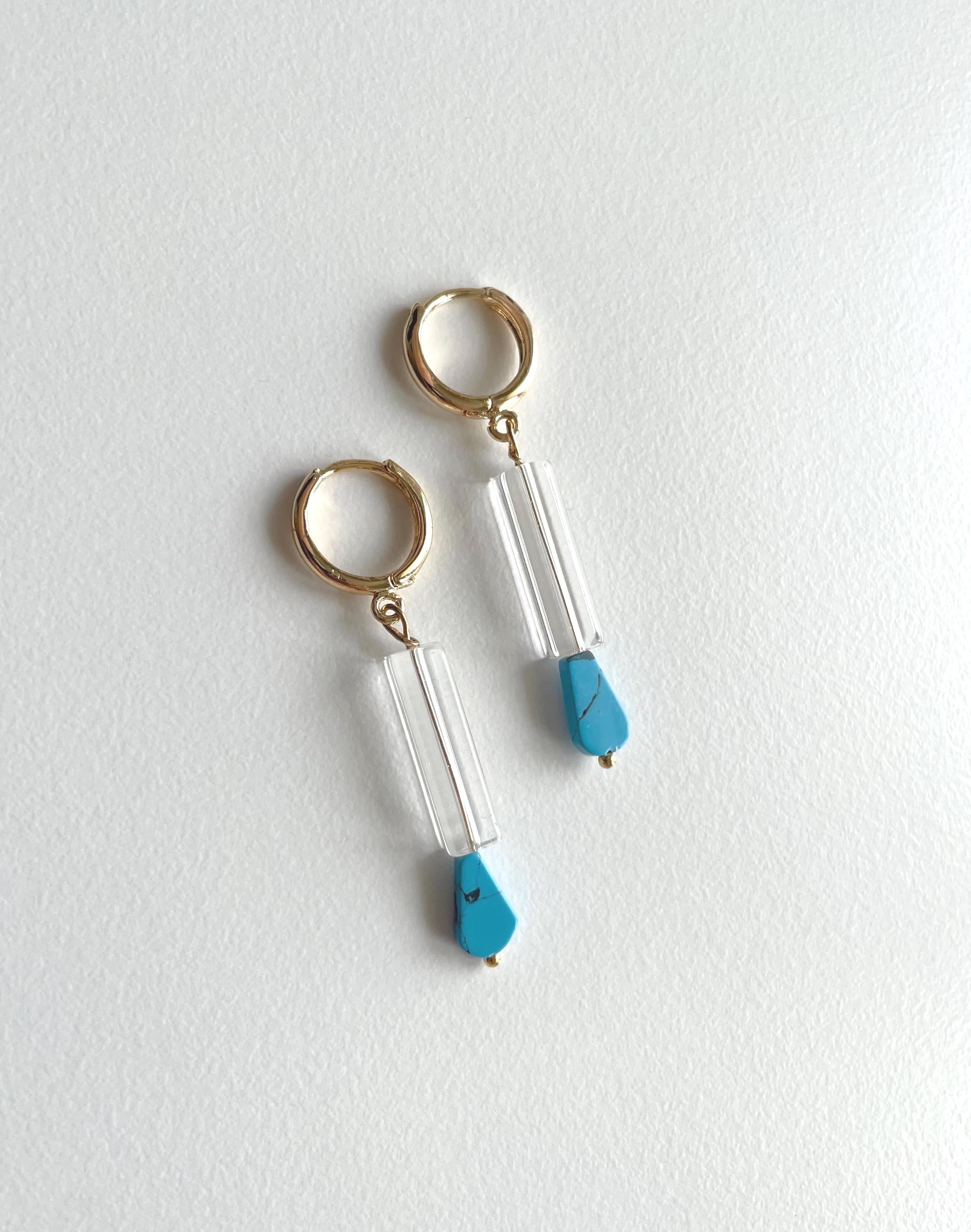 A pair of gold hoop earrings with clear cylindrical beads and bright turquoise blue teardrop charms, displayed on a textured white surface.