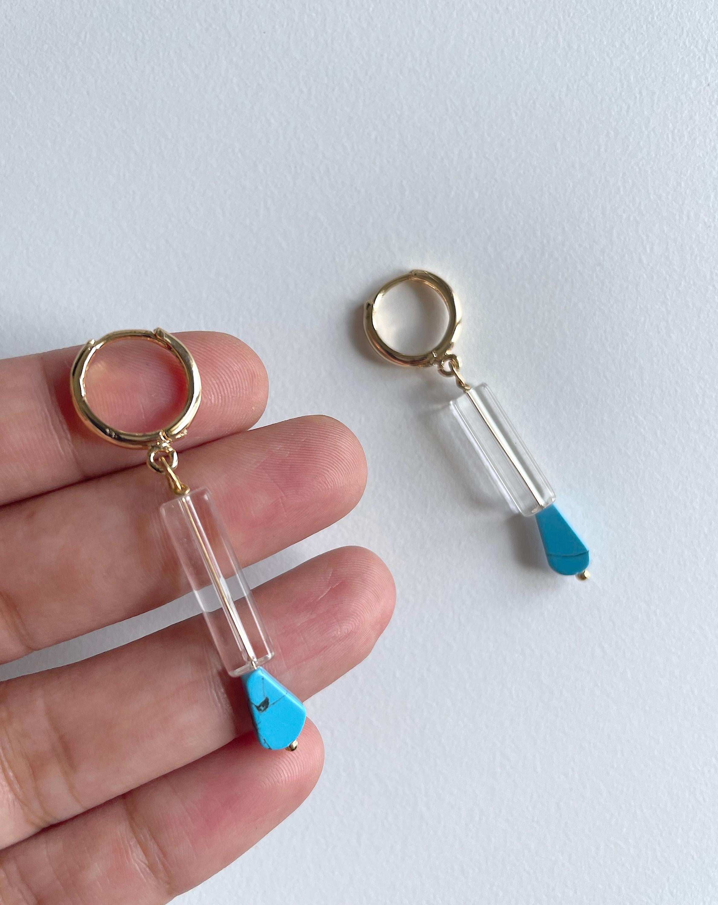 A pair of gold-tone keychains with clear cylindrical beads and bright blue teardrop charms, shown with one keychain held on a person's fingers and the other resting on a white surface.