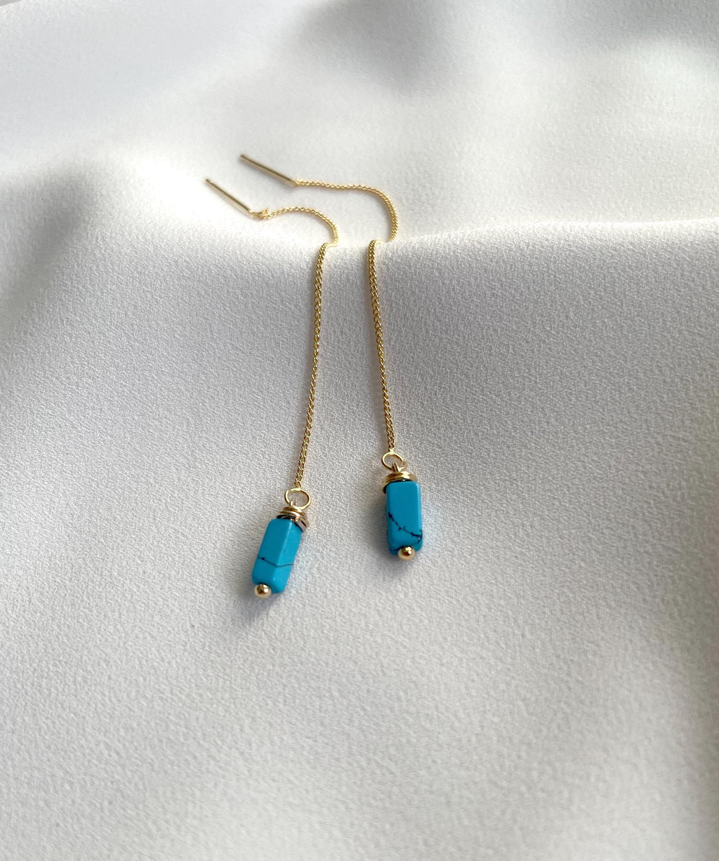 A pair of gold chain drop earrings with rectangular turquoise blue beads, displayed on a soft white fabric background.
