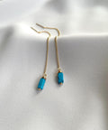 A pair of gold chain drop earrings with rectangular turquoise blue beads, displayed on a soft white fabric background.