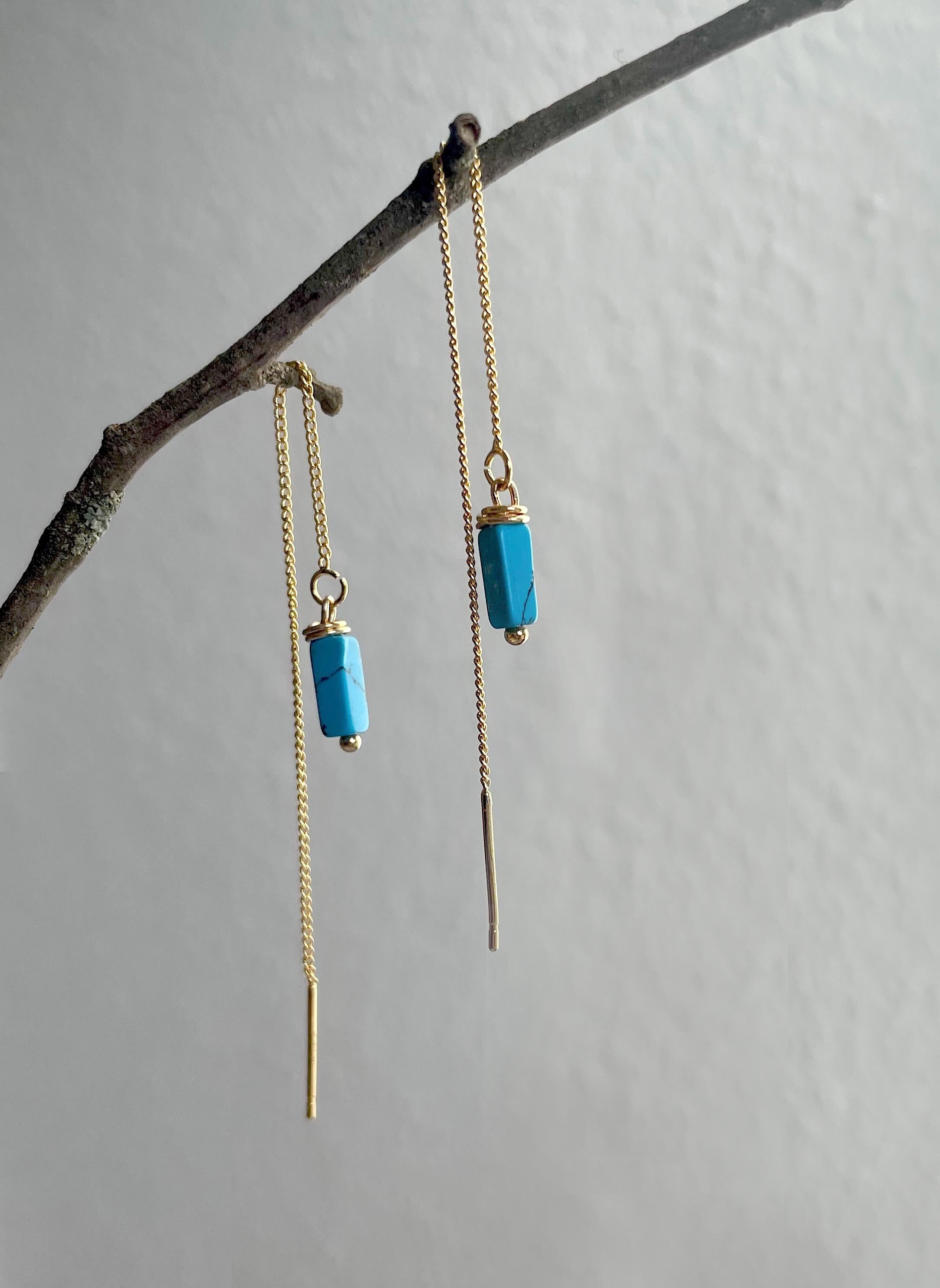 A pair of gold dangle earrings with rectangular turquoise blue beads and long drop chains, hanging from a thin branch against a light gray wall.