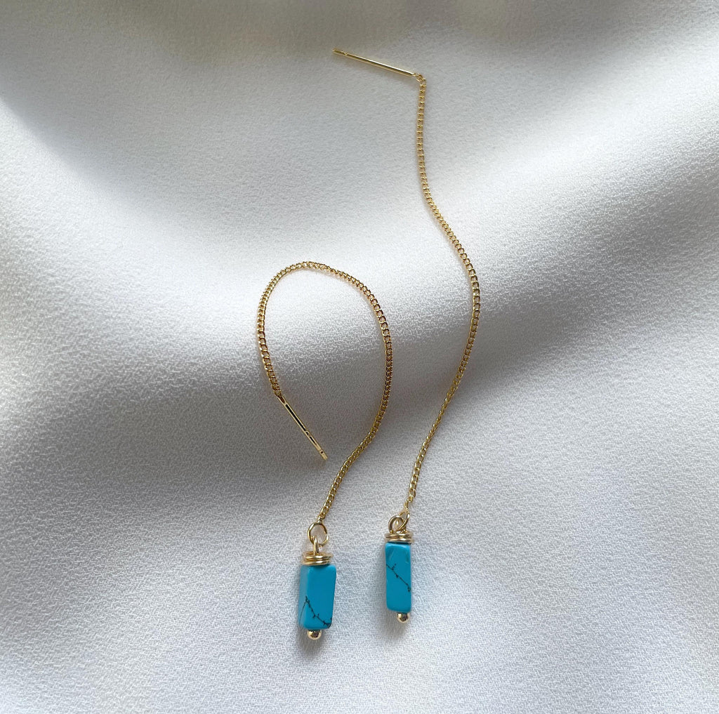 A pair of gold chain drop earrings with rectangular turquoise blue stone pendants, displayed on a textured white fabric background.
