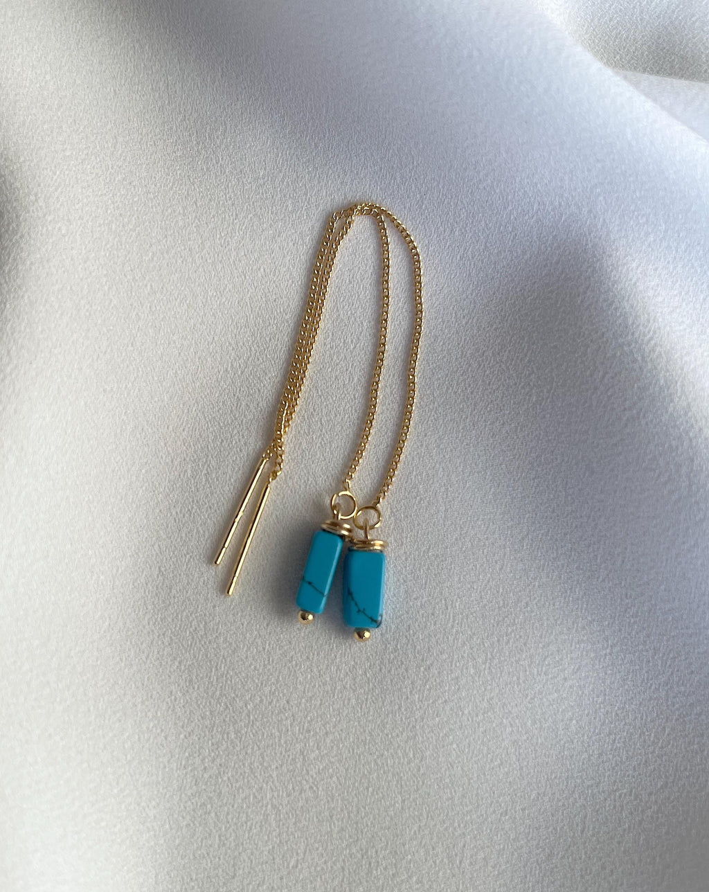 A delicate gold chain necklace featuring two cylindrical turquoise blue bead pendants with gold caps, displayed on a soft, draped white fabric background.
