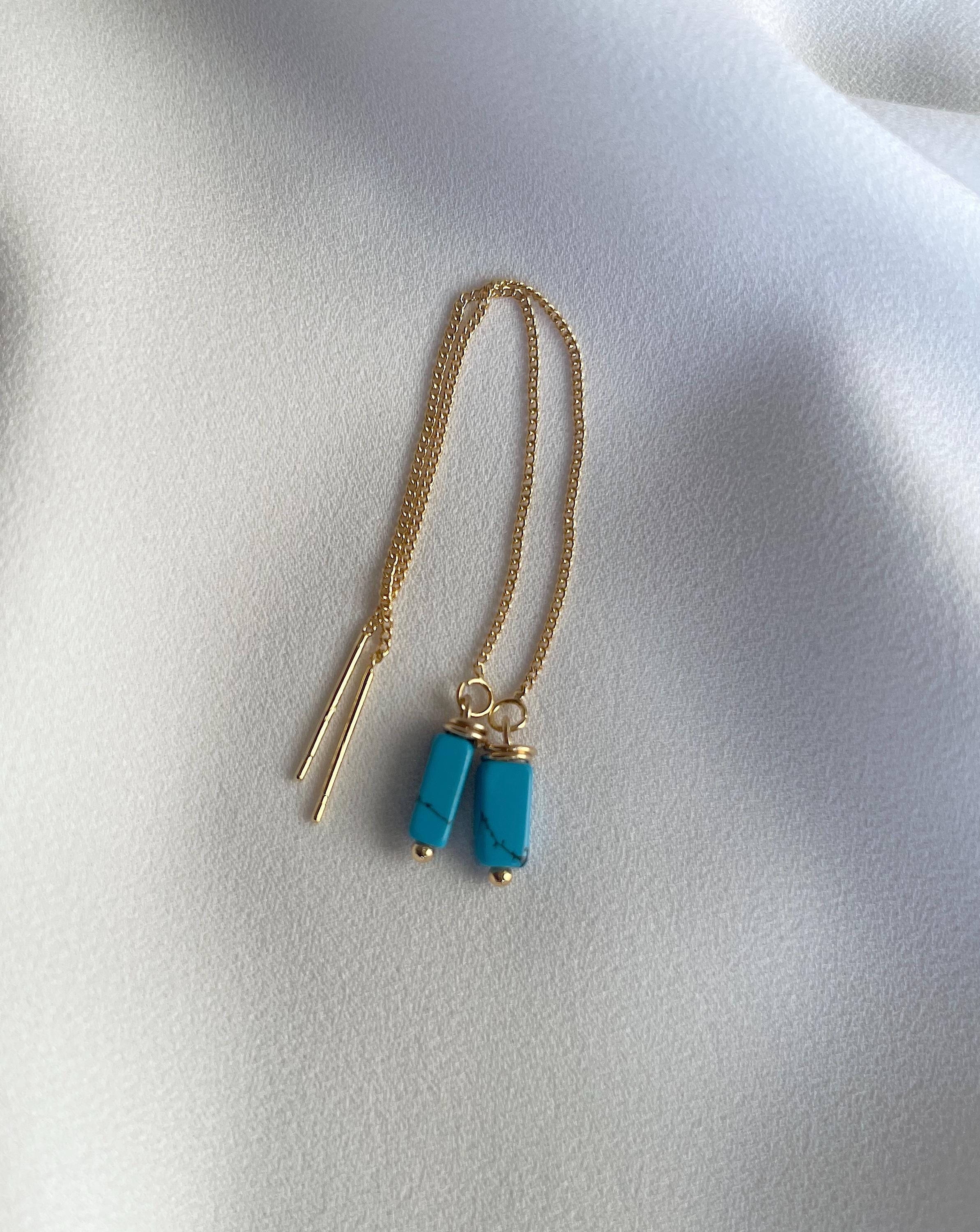 A delicate gold chain necklace featuring two cylindrical turquoise blue bead pendants with gold caps, displayed on a soft, draped white fabric background.