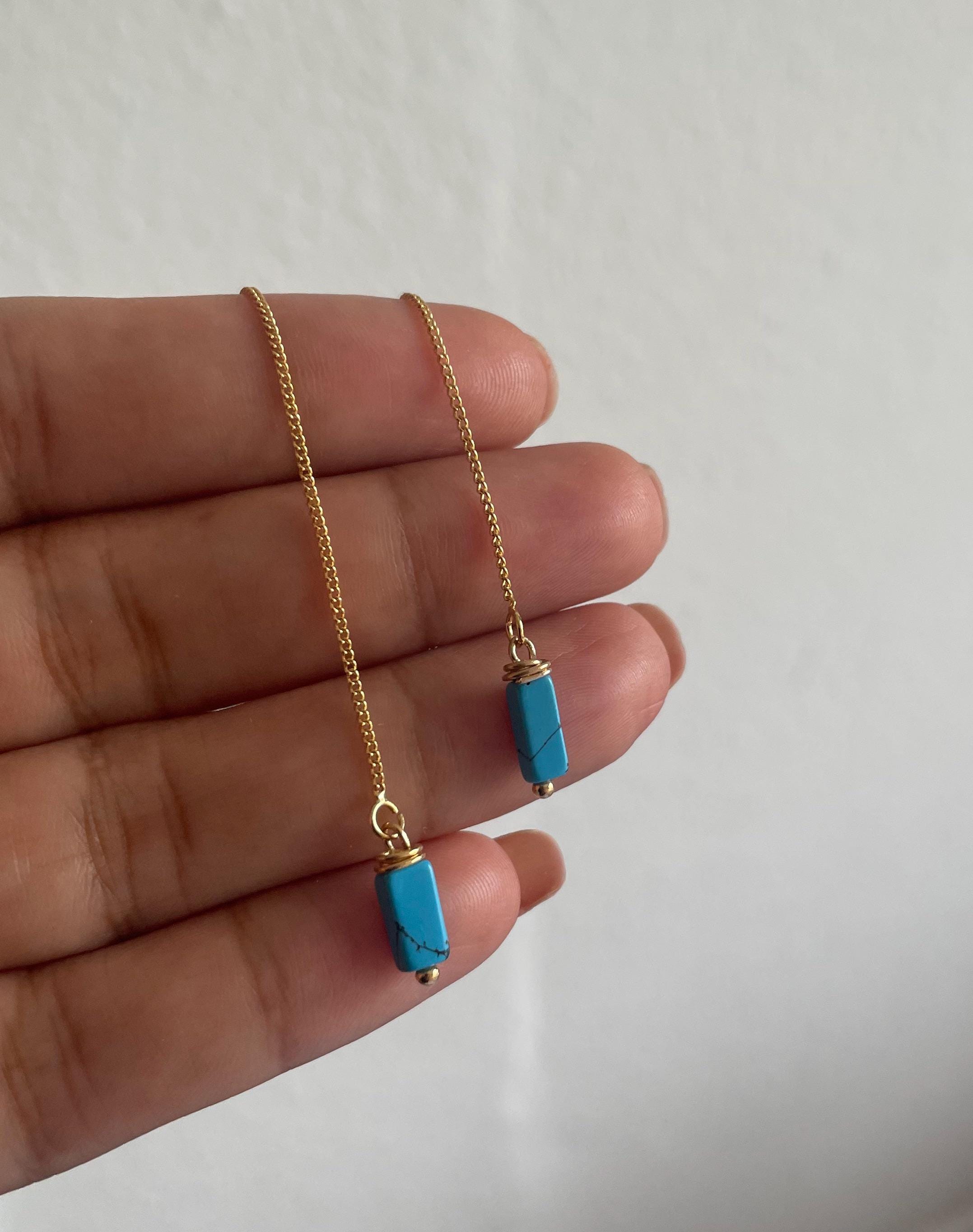 A pair of gold chain earrings with rectangular turquoise blue stone pendants, held in a person's hand against a plain light background.