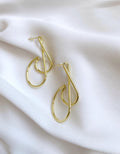 A pair of gold-toned abstract loop hoop earrings displayed on a soft, draped white fabric background.