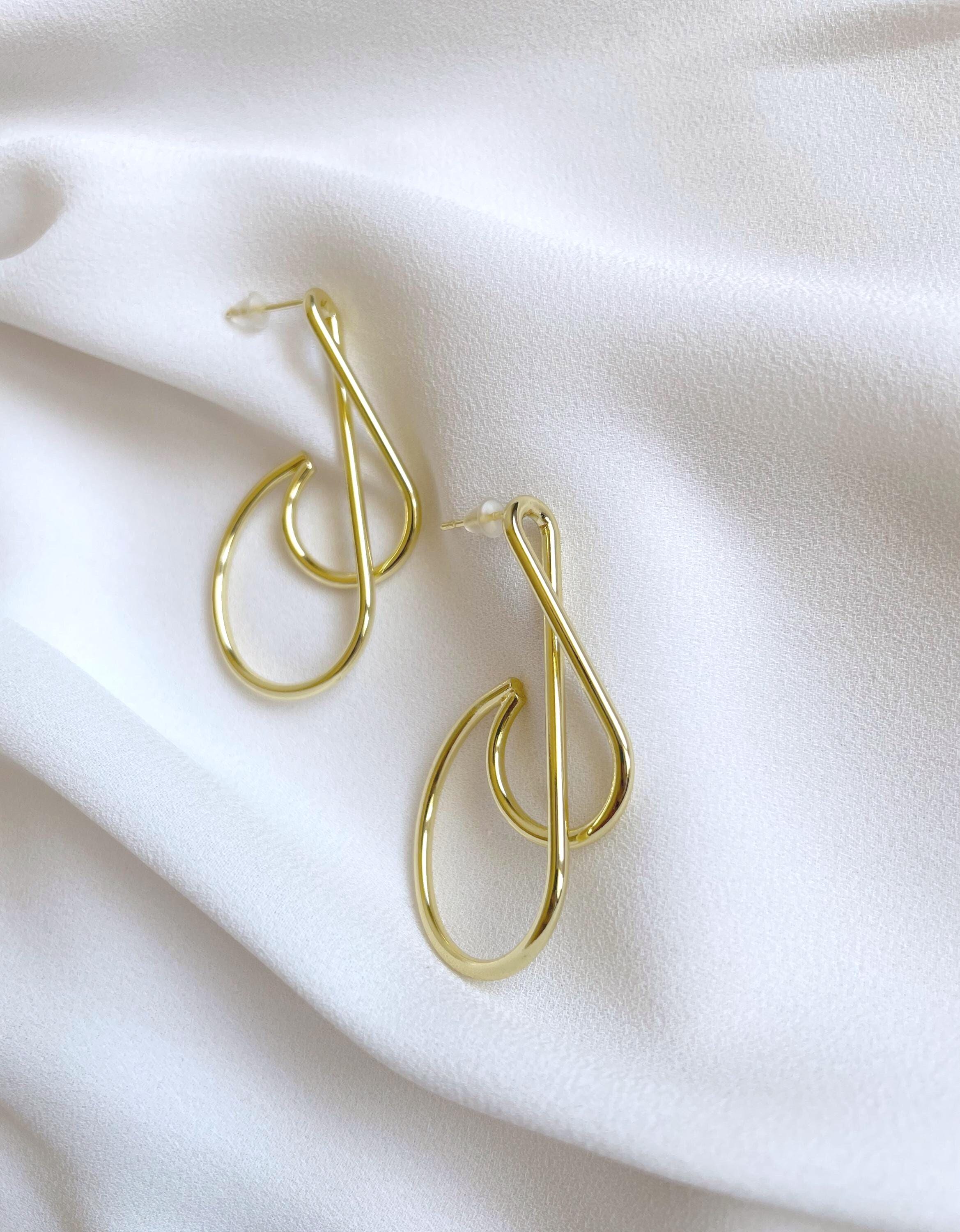 A pair of gold-toned abstract loop hoop earrings displayed on a soft, draped white fabric background.