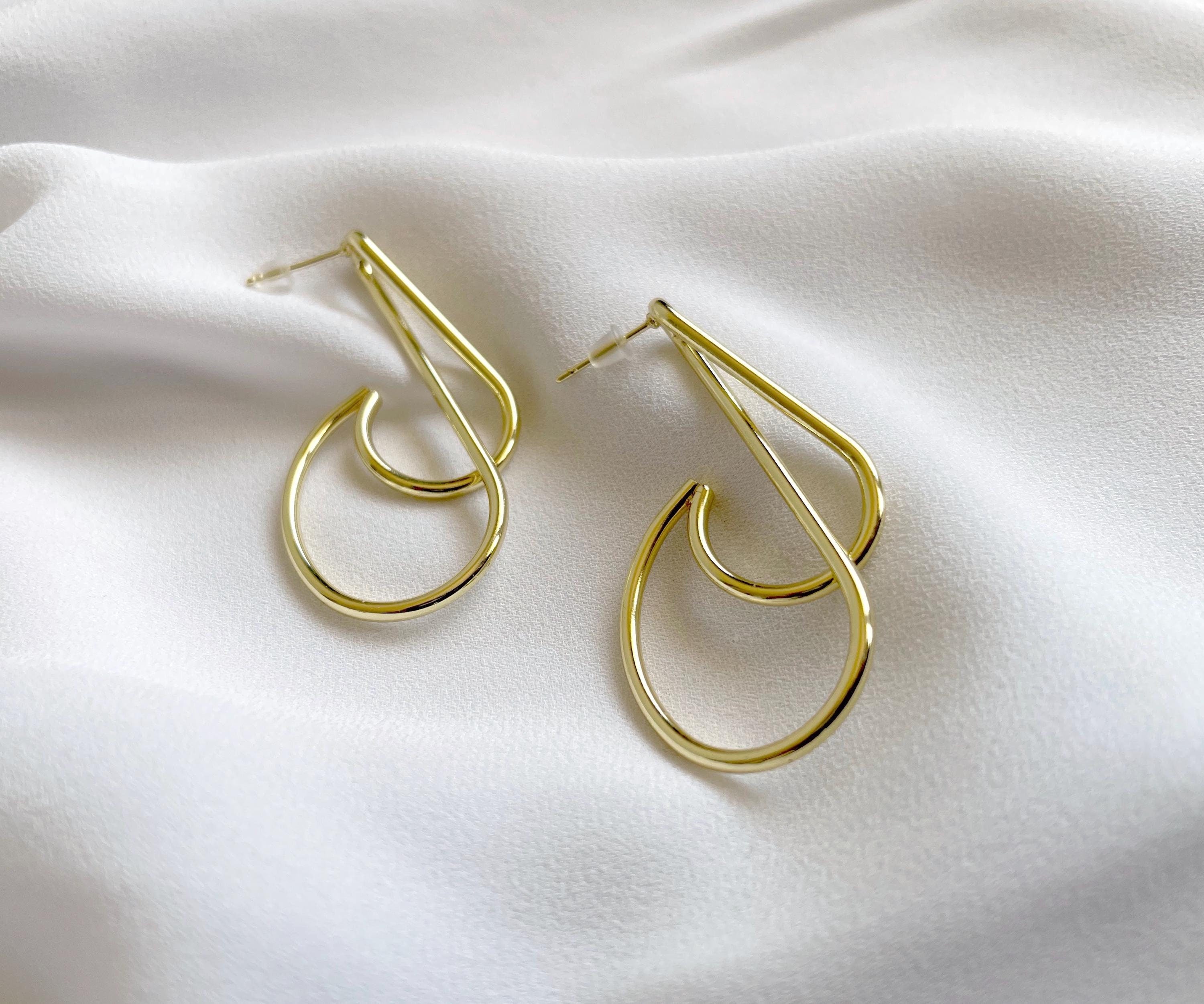 A pair of gold-toned abstract loop hoop earrings resting on a soft, draped white fabric background.