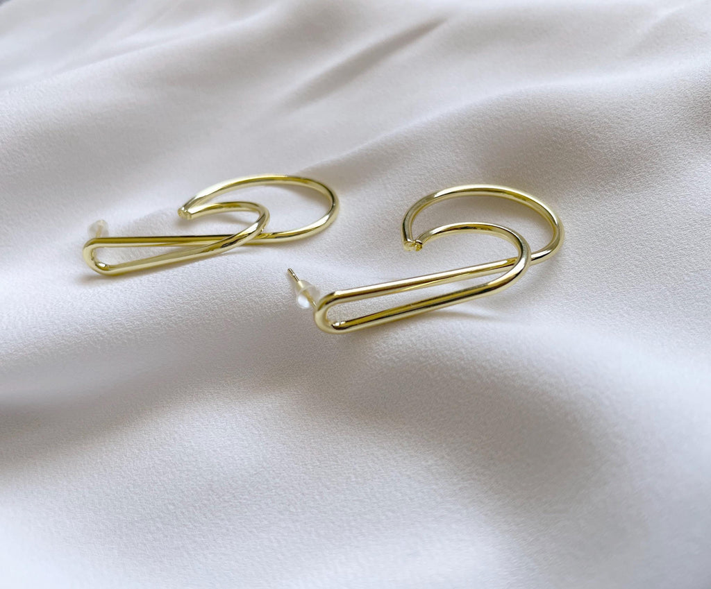 A pair of gold wire hoop earrings with a minimalist, open-ended design, displayed on a soft, draped white fabric background.