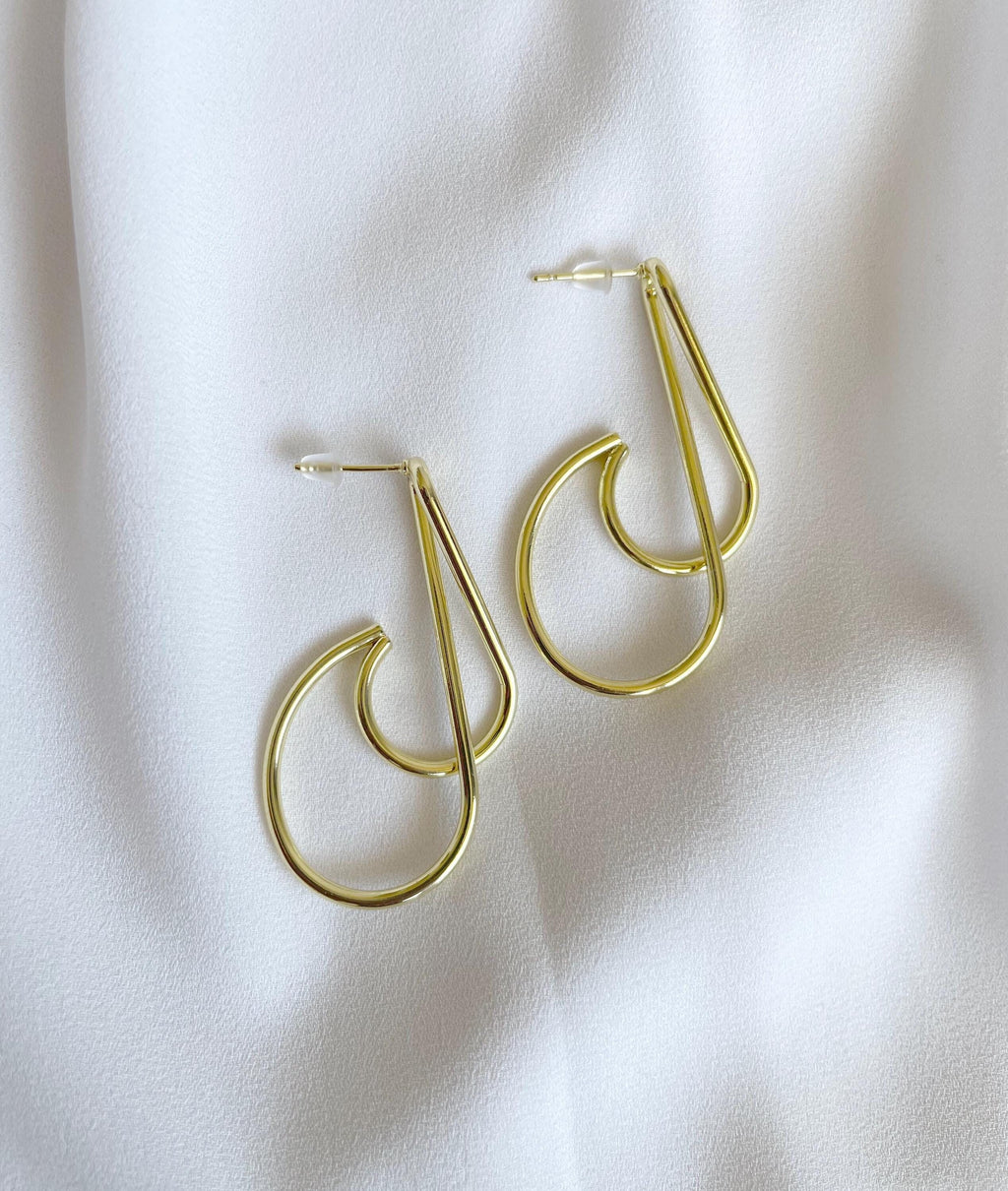 A pair of gold-toned geometric hoop earrings with a double-loop design, displayed on a soft, white textured fabric background.