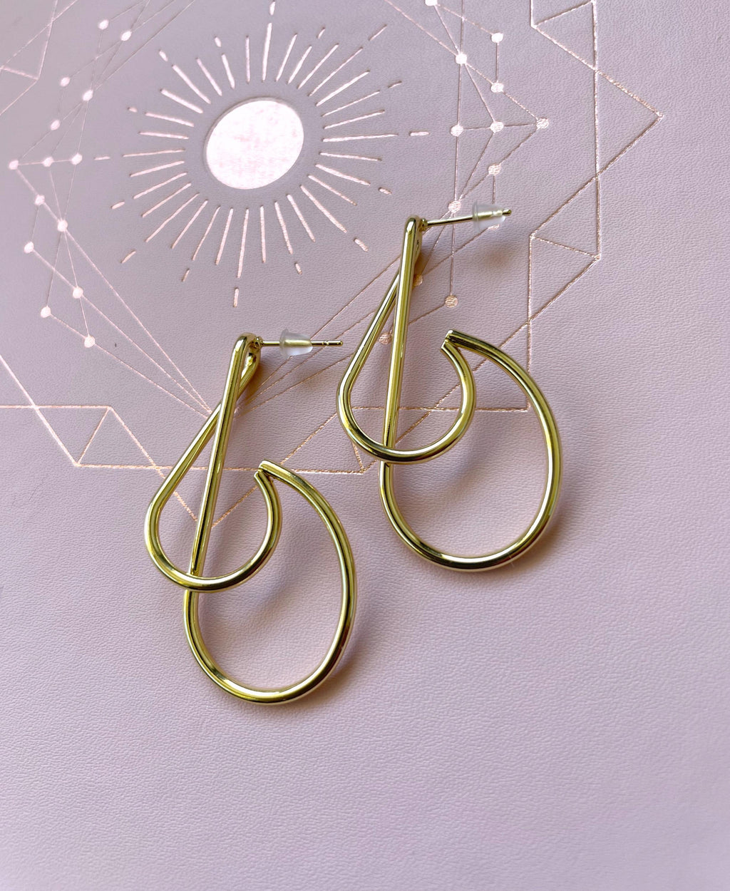 A pair of gold-toned open hoop earrings with a teardrop-like curved design, displayed on a light lavender surface with a geometric sun illustration in the background.