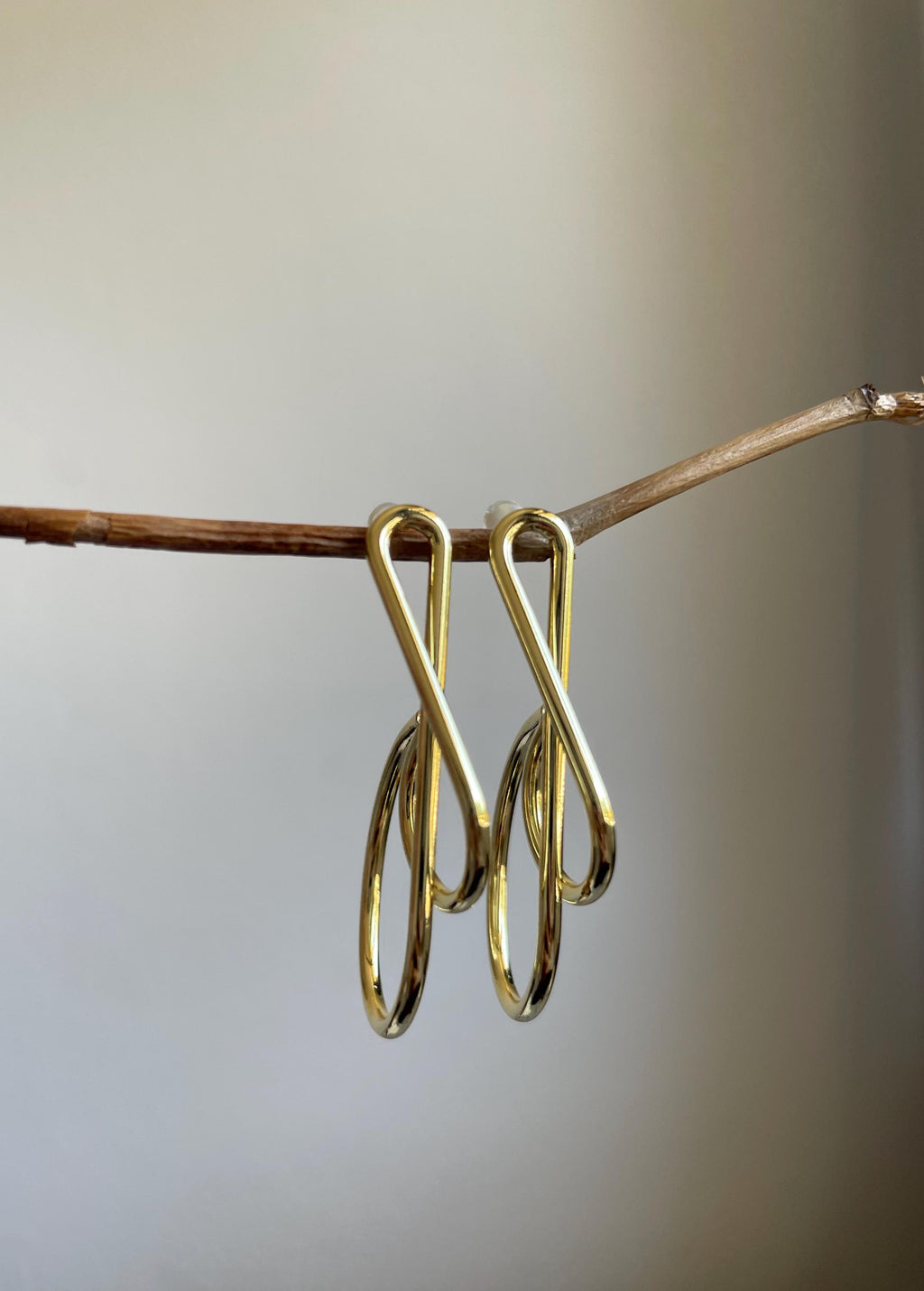 A pair of polished gold-toned metal earrings with an infinity-shaped top and dangling oval drops, hanging from a thin brown twig against a soft neutral gray background.
