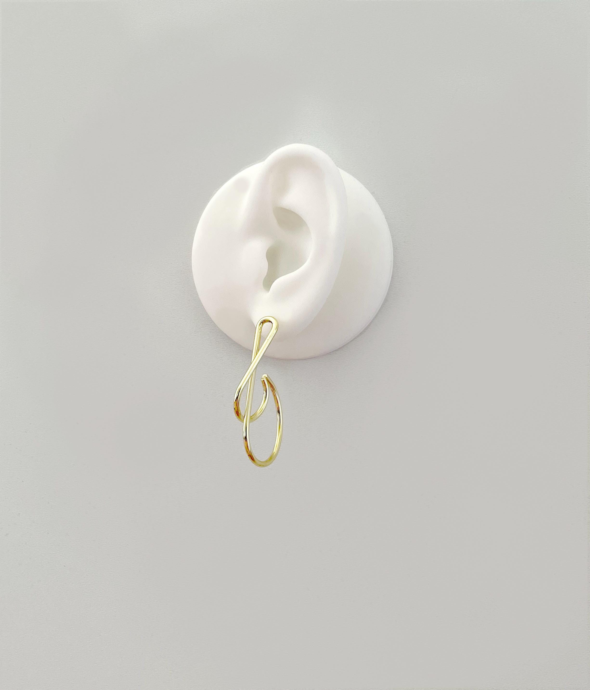 A gold-colored hoop earring with a twisted, organic loop design, shown hanging from a white ear-shaped display against a solid light gray background.