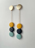 A pair of long drop earrings with matte circular discs in yellow, charcoal gray, and light mint green, suspended from gold-toned chains and a gold-toned bar, shown against a plain off-white wall.