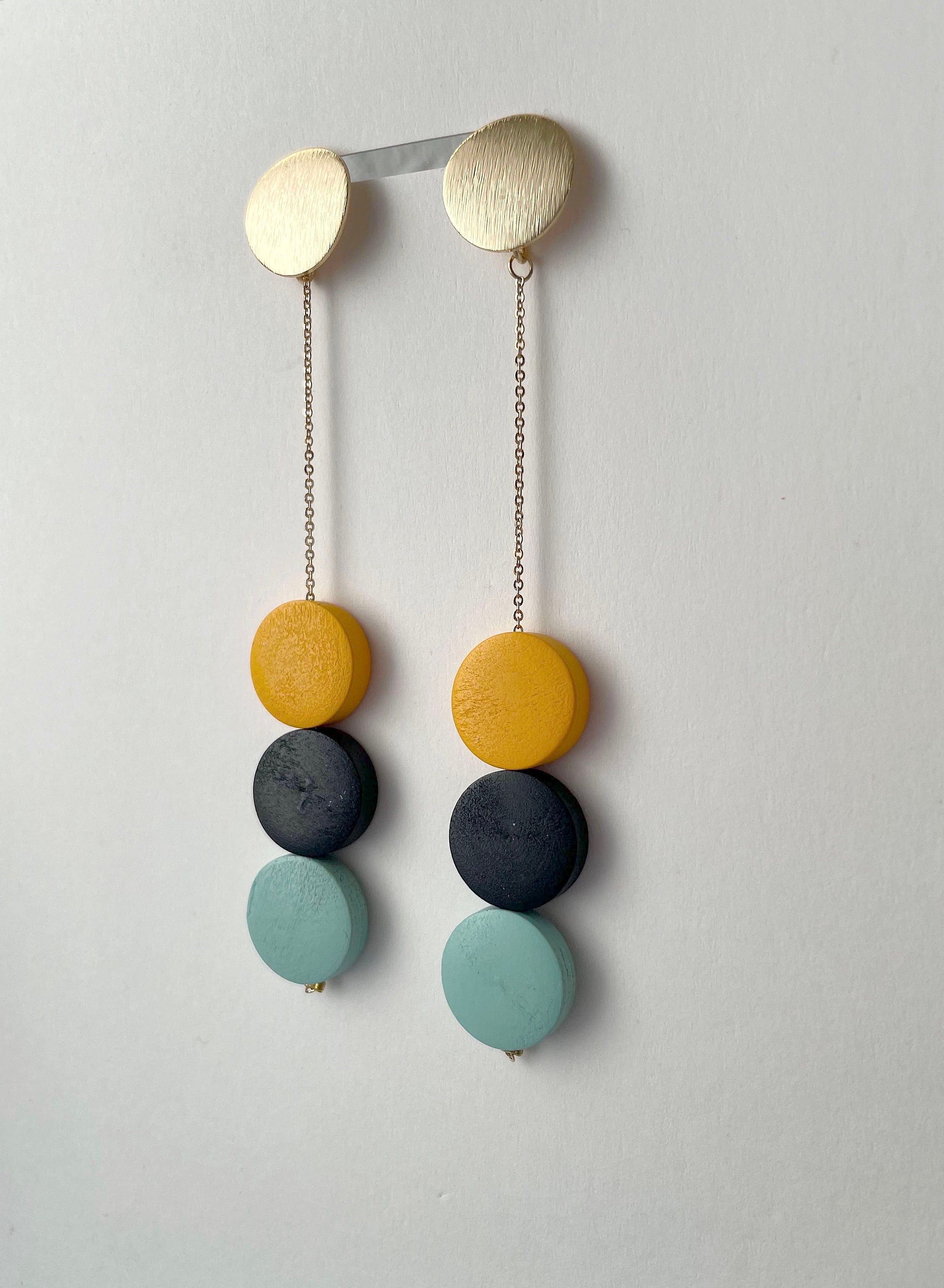 A pair of long drop earrings with matte circular discs in yellow, charcoal gray, and light mint green, suspended from gold-toned chains and a gold-toned bar, shown against a plain off-white wall.