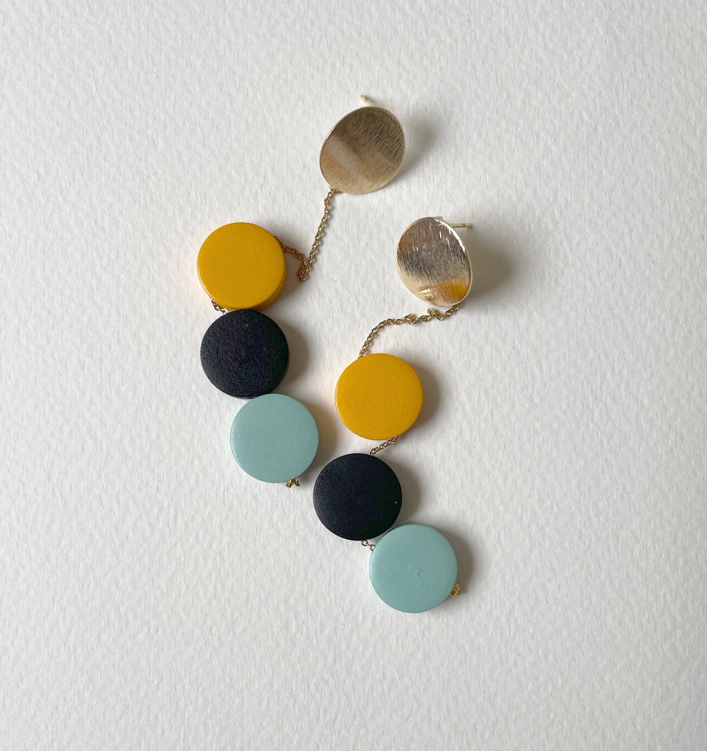 A pair of drop earrings with alternating round wooden discs in mustard yellow, black, and light blue, suspended from gold-toned chains with flat gold disc studs, displayed on a textured white background.