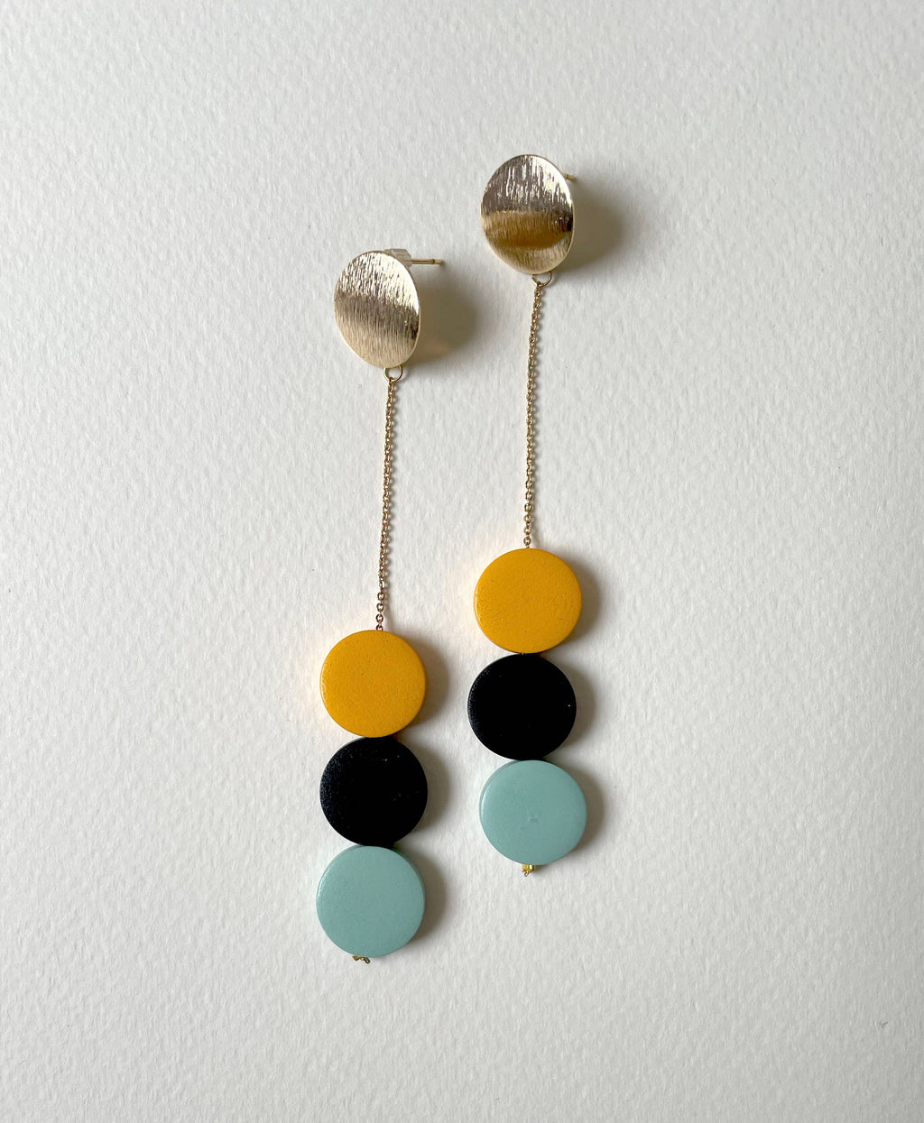 A pair of modern dangle earrings with textured gold-toned studs, hanging chains, and three stacked circular beads in yellow, black, and light blue on a textured off-white background.