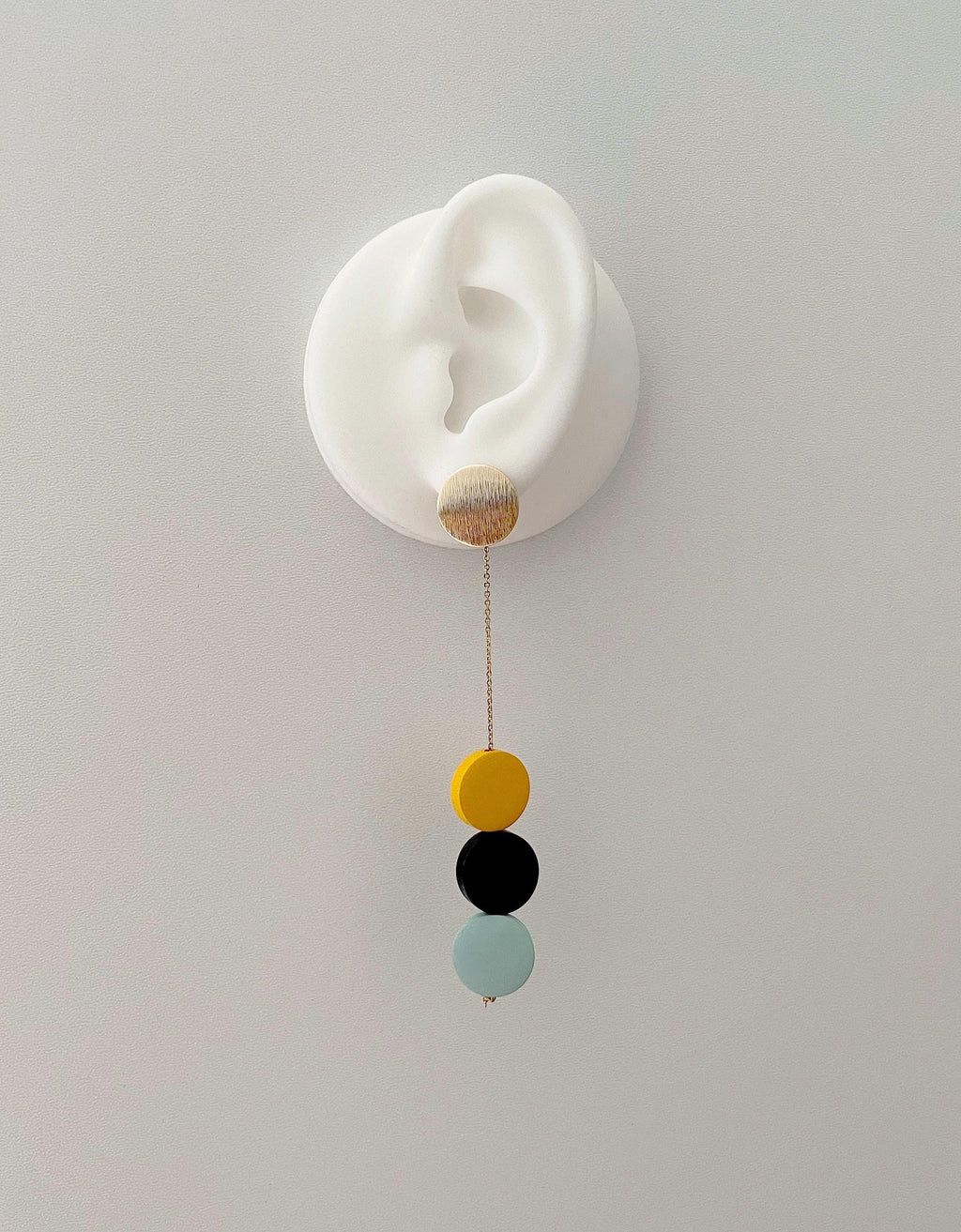 A white silicone ear-shaped wall hook with a dangling gold-tone chain and three round beads in yellow, black, and pale mint, shown against a plain light gray wall.