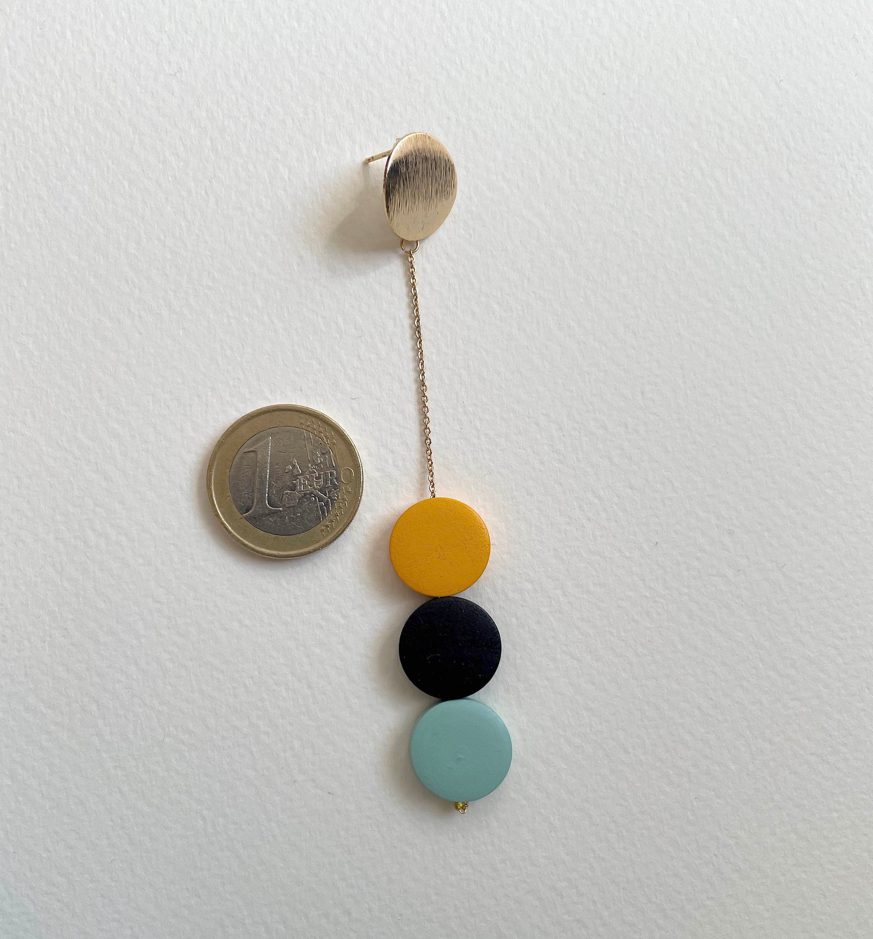 A minimalist gold-tone stud earring with a long chain and four hanging circular beads in mustard yellow, black, and light blue, shown next to a 1 Euro coin for scale on a white background.