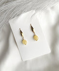 A pair of gold drop earrings with oval yellow beads, displayed on a white card over a white fabric background with soft gray decorative fibers.