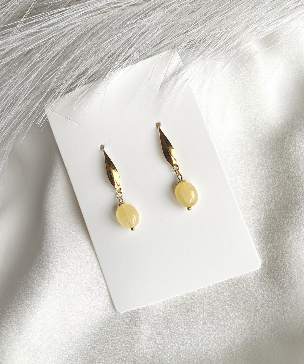 A pair of gold plated drop earrings with oval yellow natural calcite beads, displayed on a white card over a white fabric background with soft gray decorative fibers.