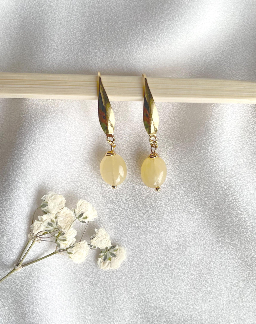 A pair of gold drop earrings with teardrop-shaped gold charms and round pale yellow beads, displayed on a light wooden stick against a white fabric background with small white flowers in the corner.