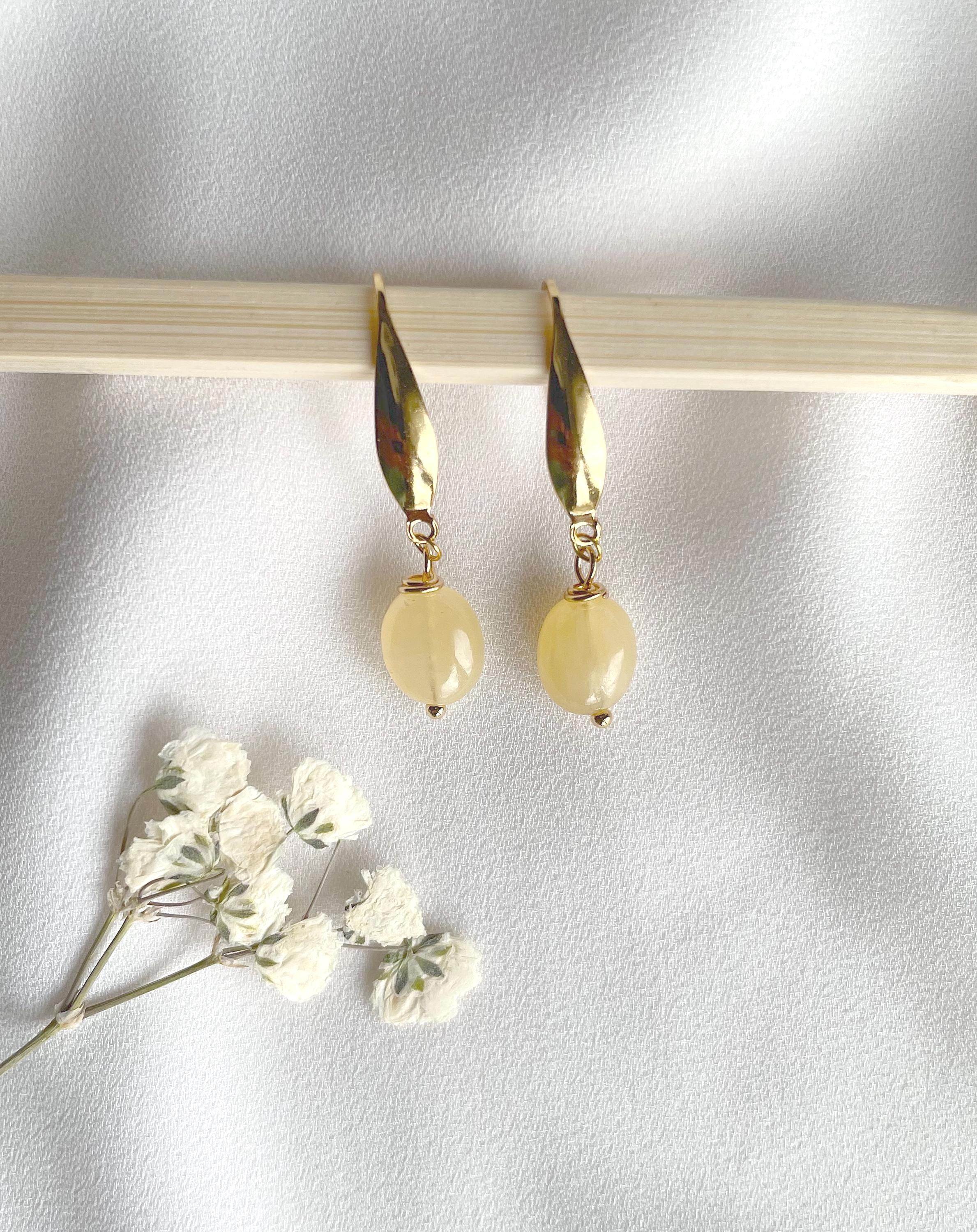 A pair of gold drop earrings with teardrop-shaped gold charms and round pale yellow beads, displayed on a light wooden stick against a white fabric background with small white flowers in the corner.