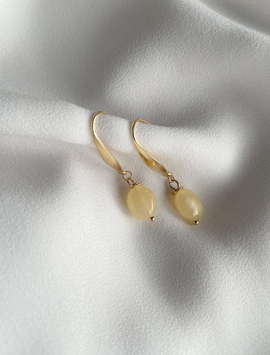A pair of gold drop earrings with smooth, oval pale yellow beads, displayed on a textured white fabric background.