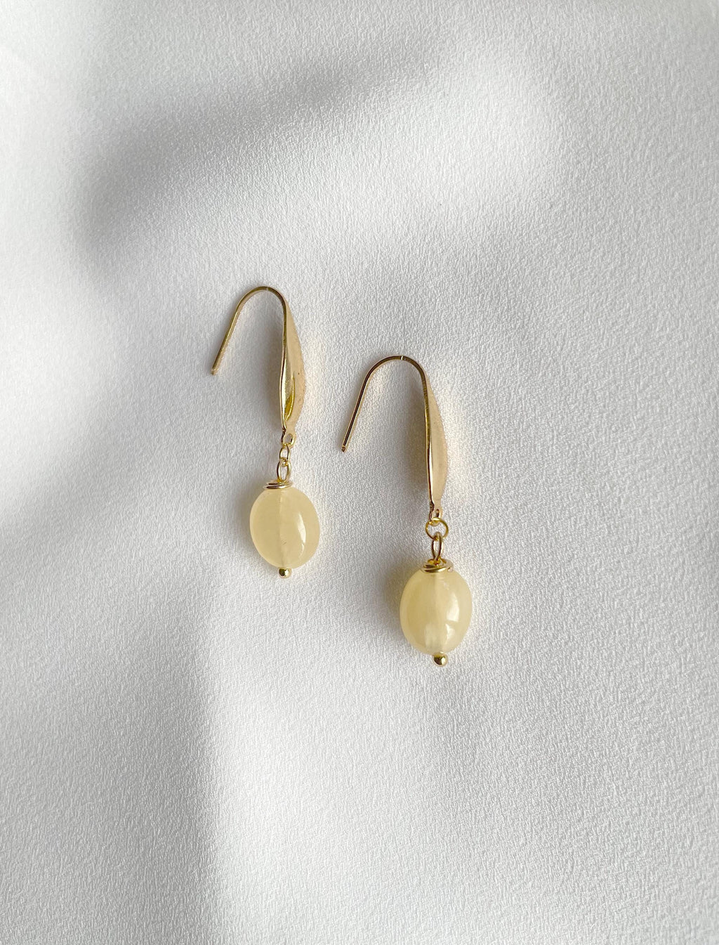 A pair of gold-toned dangle earrings with smooth, oval pale yellow beads, displayed on a textured white surface with soft shadows.