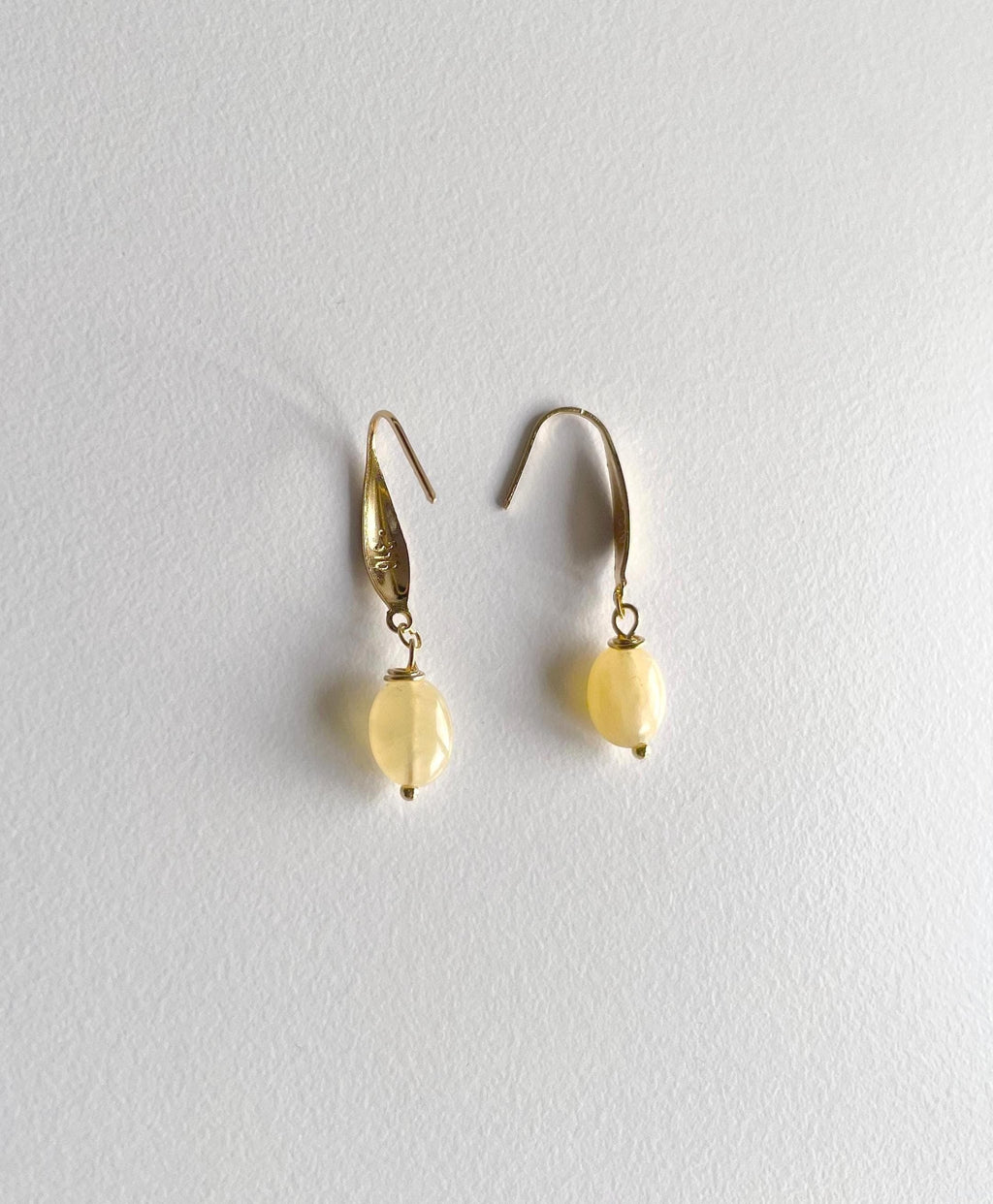 A pair of gold drop earrings with small, oval, pale yellow beads hanging from simple gold fishhook wires, displayed against a textured off-white background.