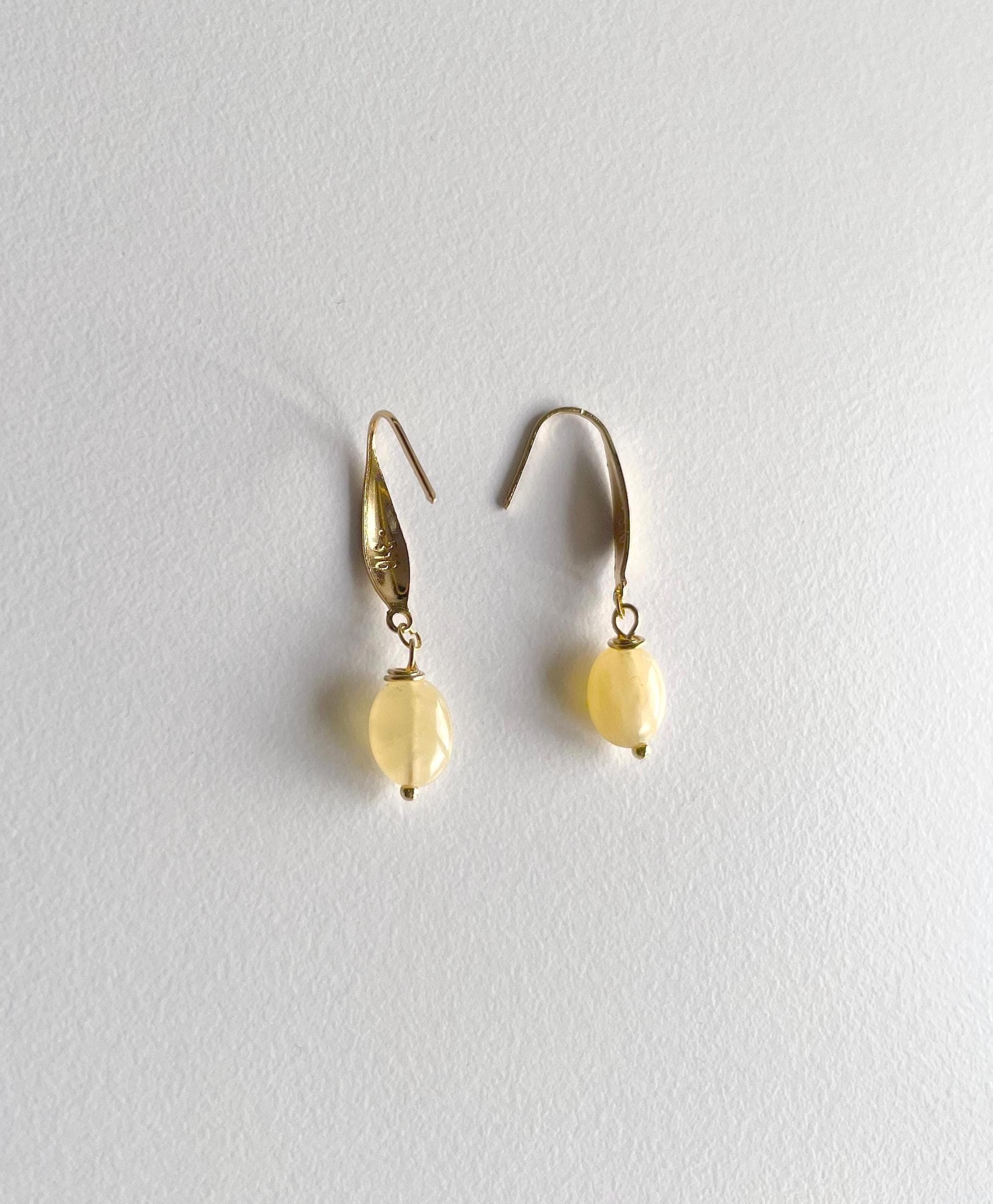 A pair of gold drop earrings with small, oval, pale yellow beads hanging from simple gold fishhook wires, displayed against a textured off-white background.