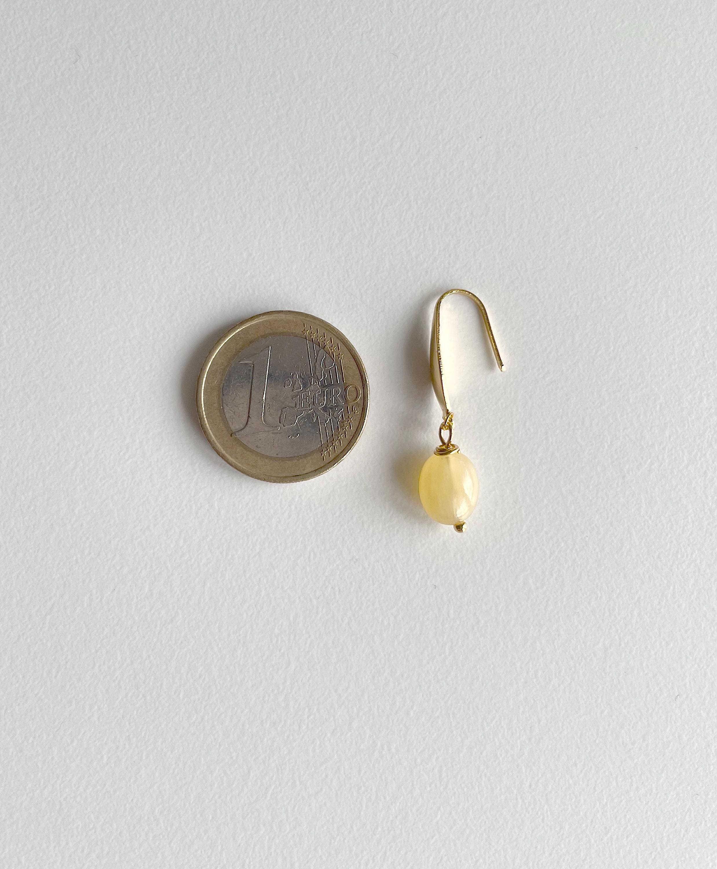 A single small gold dangle earring with a pale yellow bead drop, shown next to a euro coin for scale on a light gray background.