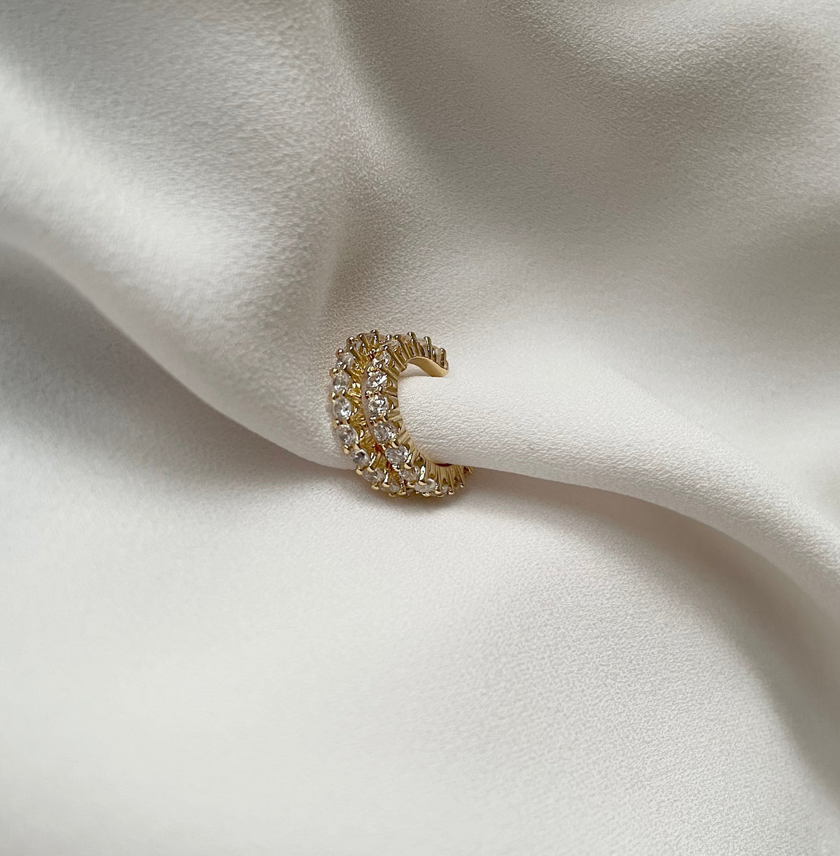 A pair of gold plated crystal cuff earrings paved with small cubic zirconia clear stones, resting on draped white satin fabric.