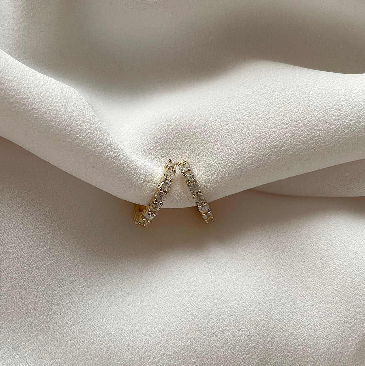 A pair of delicate gold plated ear cuffs adorned with small clear pavé stones, resting on draped white satin fabric.