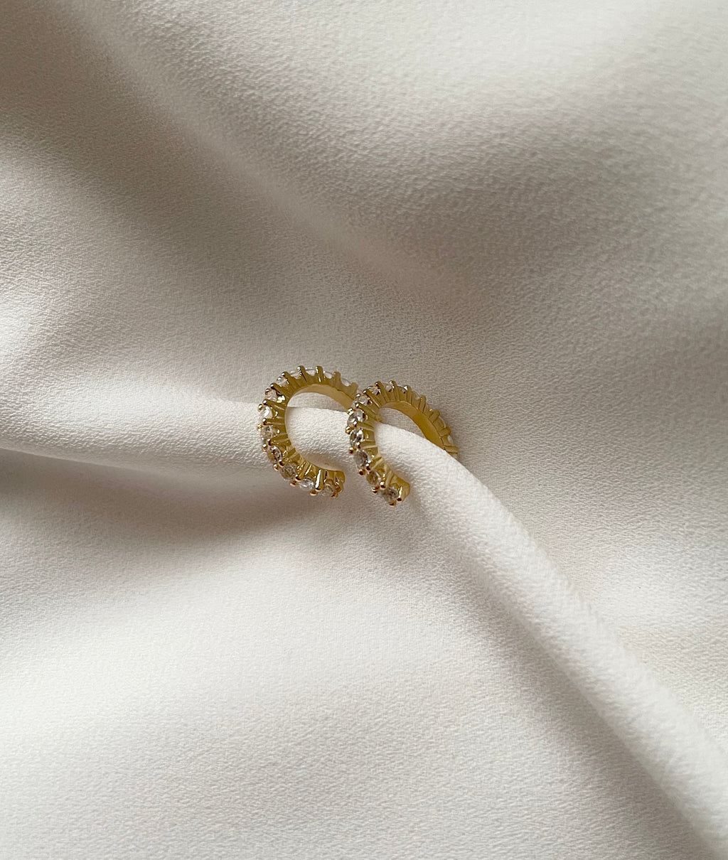 A pair of gold plated cuff earrings with small clear stones, resting on draped white fabric.