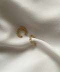 A pair of gold cuff earrings with clear rectangular crystals, displayed on a draped white fabric background.