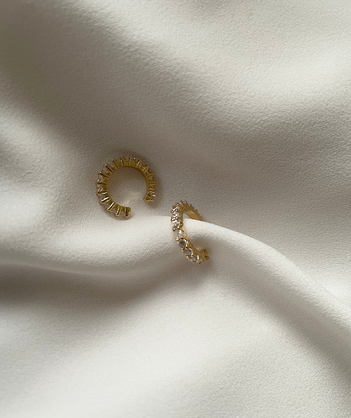 A pair of gold cuff earrings with clear rectangular crystals, displayed on a draped white fabric background.