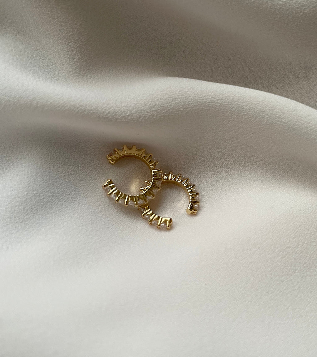 A pair of gold cuff earrings with textured, ridged edges, displayed on a soft, draped white fabric background.