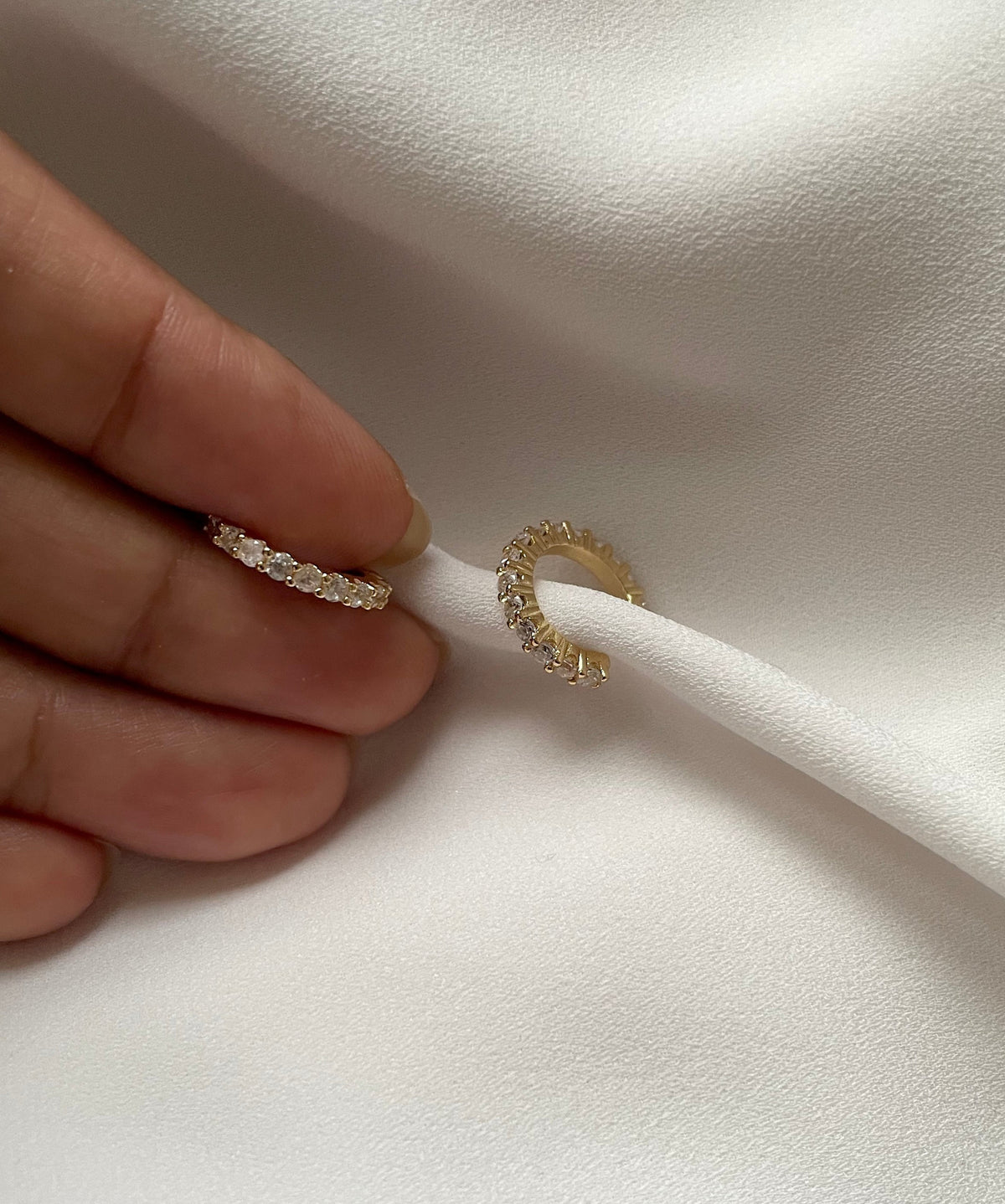 A close-up of a hand holding a thin gold ear cuff with clear pavé stones, next to another gold cuff earring on a white textured fabric background.