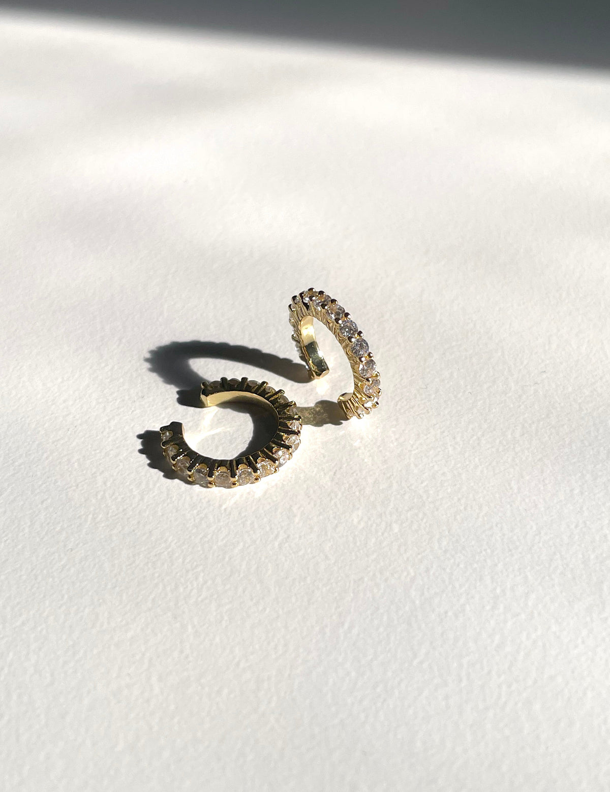 A pair of gold cuff earrings adorned with small clear crystals, resting on a textured white surface with a soft shadow.