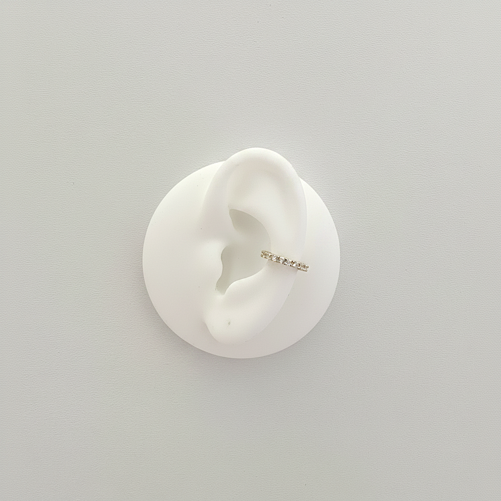 A delicate silver ear cuff with small clear stones, displayed on a white ear-shaped stand against a light gray background.