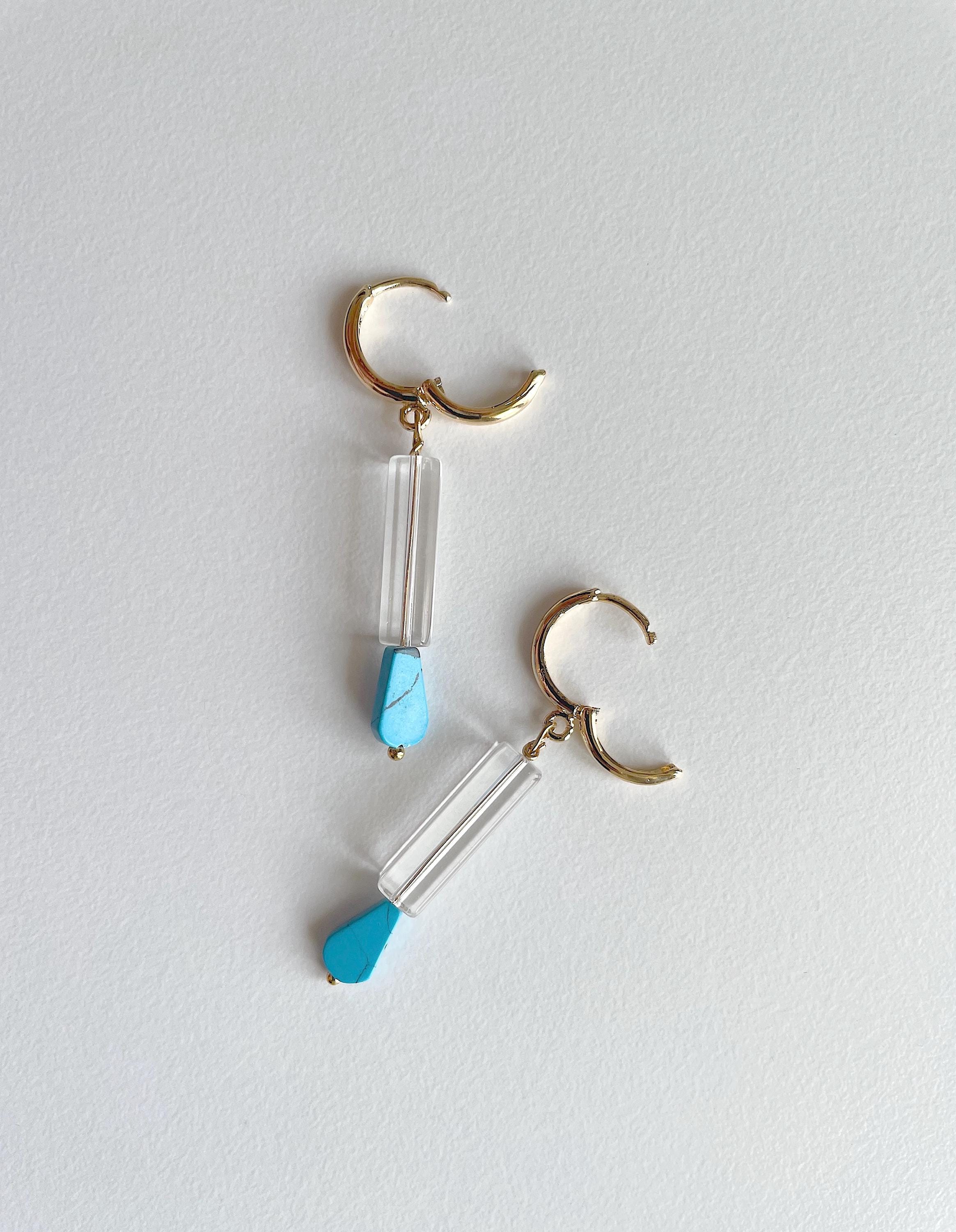 A pair of gold hoop dangle earrings with clear glass cylinder drops and small light blue teardrop-shaped accents, displayed on a light gray textured background.