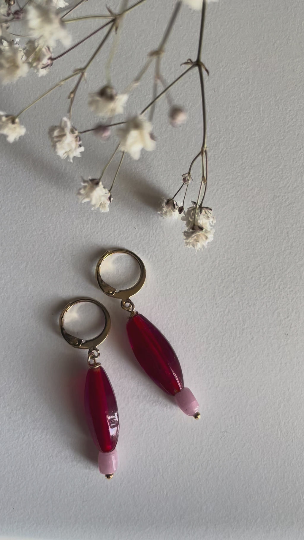 Red Earrings, A pair of gold plated huggie hoops with red elongated glass bead charms with small pastel pink beads shown on a white surface with natural sunlight.